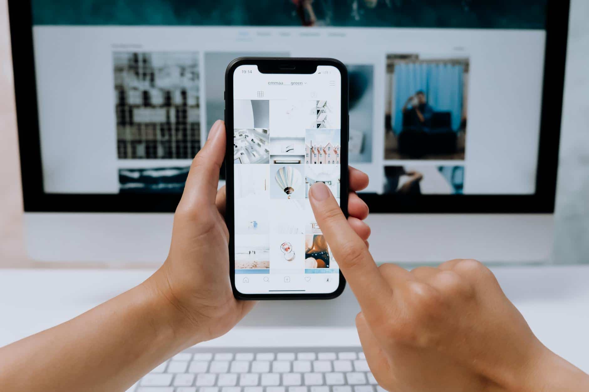 Hands holding a smartphone while browsing a social media app in front of a desktop computer.